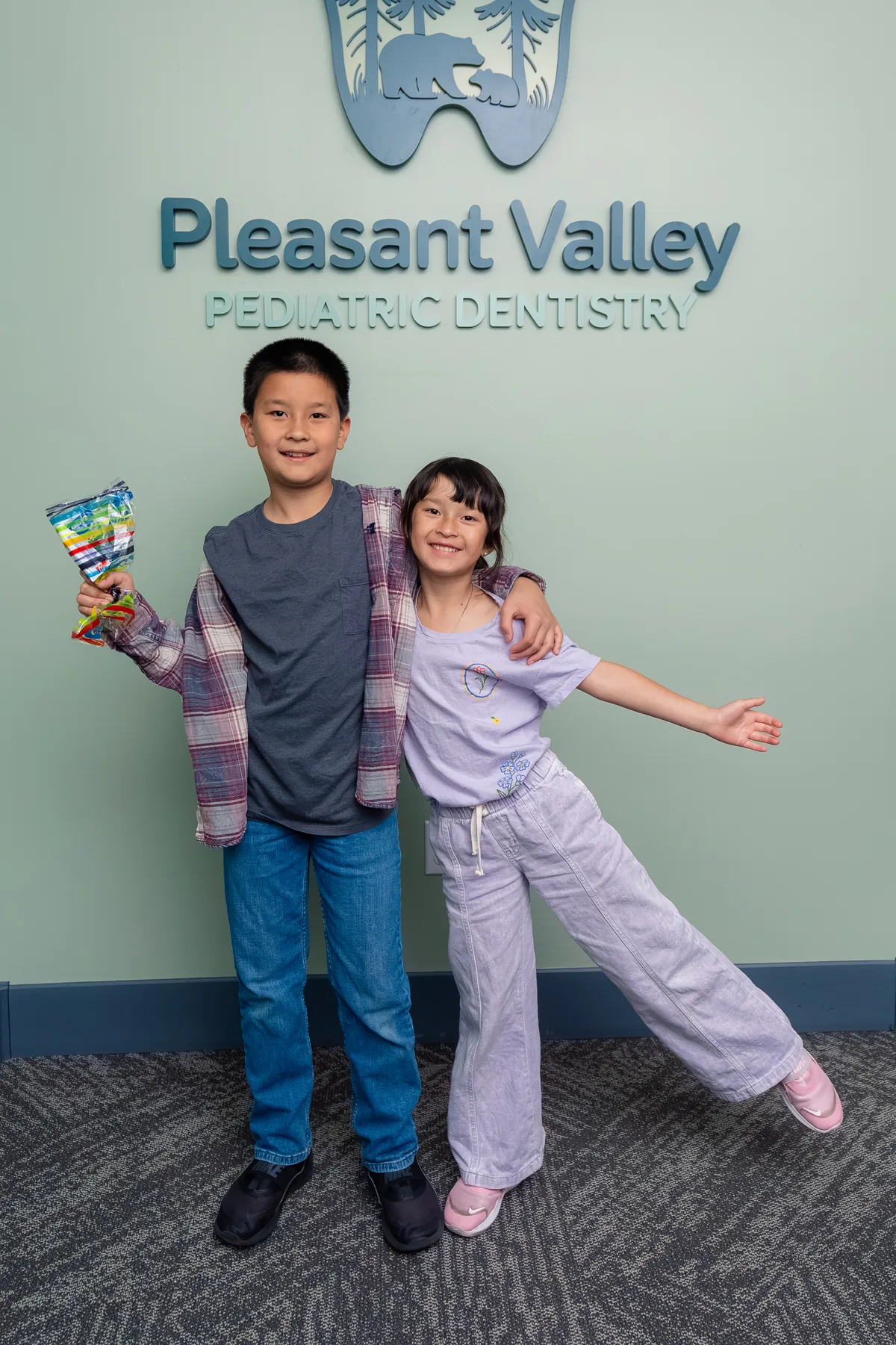 Two young patients smiling in front of the Pleasant Valley Pediatric Dentistry sign